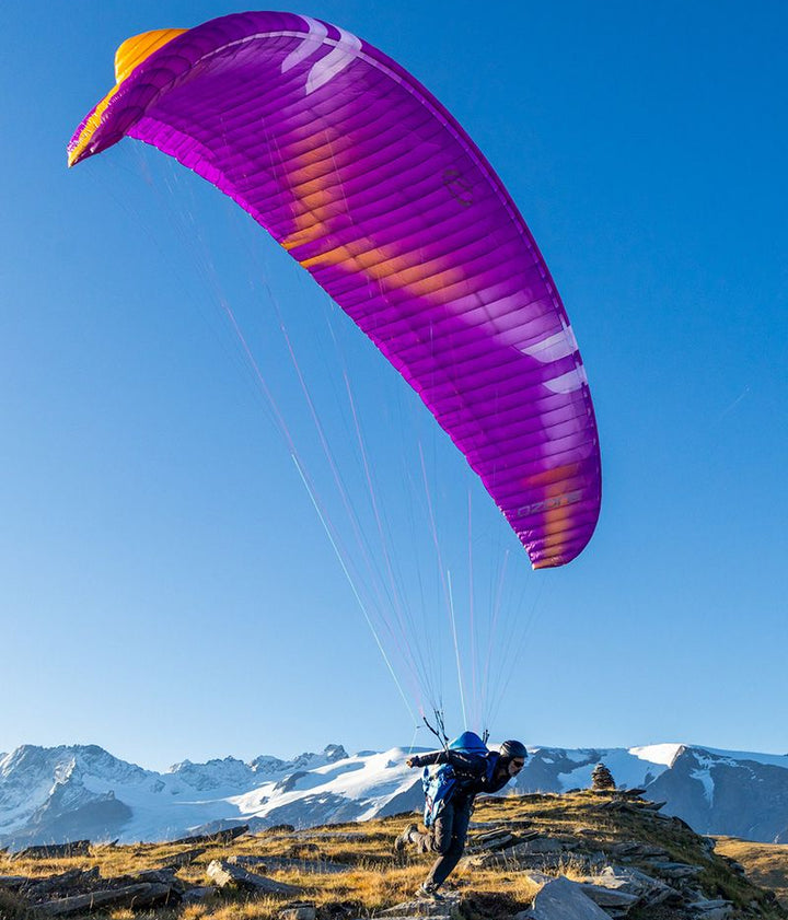 Person paragliding with a purple and yellow parachute against a mountainous landscape.